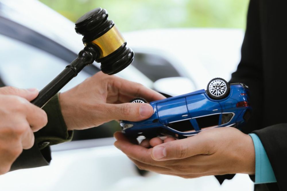A person holding a gavel prepares to finalize a transaction, handing over a blue toy car to another individual, set in an outdoor environment with a blurred background of vehicles.