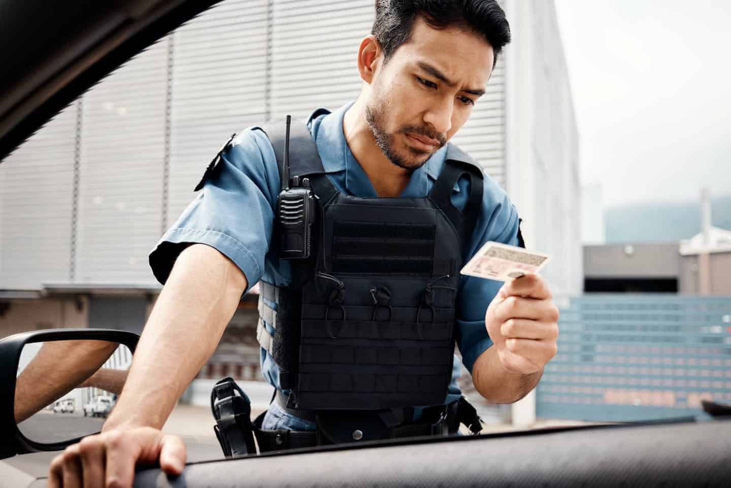 A police officer wearing a bulletproof vest is examining a piece of currency near an open car door with buildings in the background.