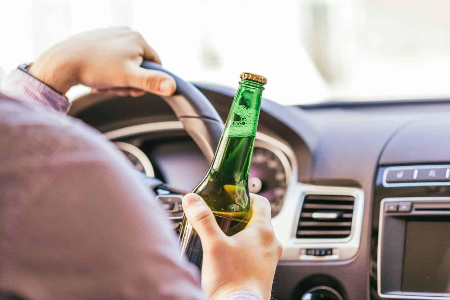 A person holds a beer while steering a car, suggesting drinking and driving, with focus on the driver's hand and bottle, blurring the dashboard and road ahead.