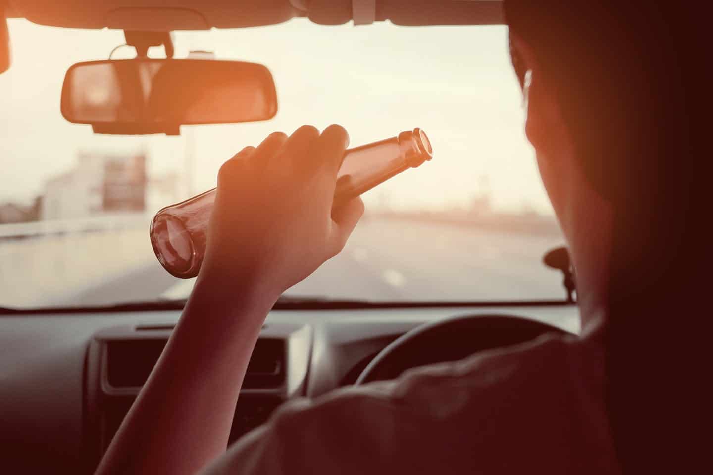 A hand holds a glass bottle inside a moving car, viewed from the backseat, with the road visible through the windshield, suggesting potential distracted driving.