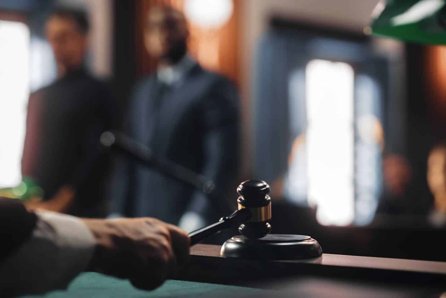 A judge's gavel rests on a sound block, with a hand poised to strike. In the blurred background, two individuals stand in a dimly lit courtroom.