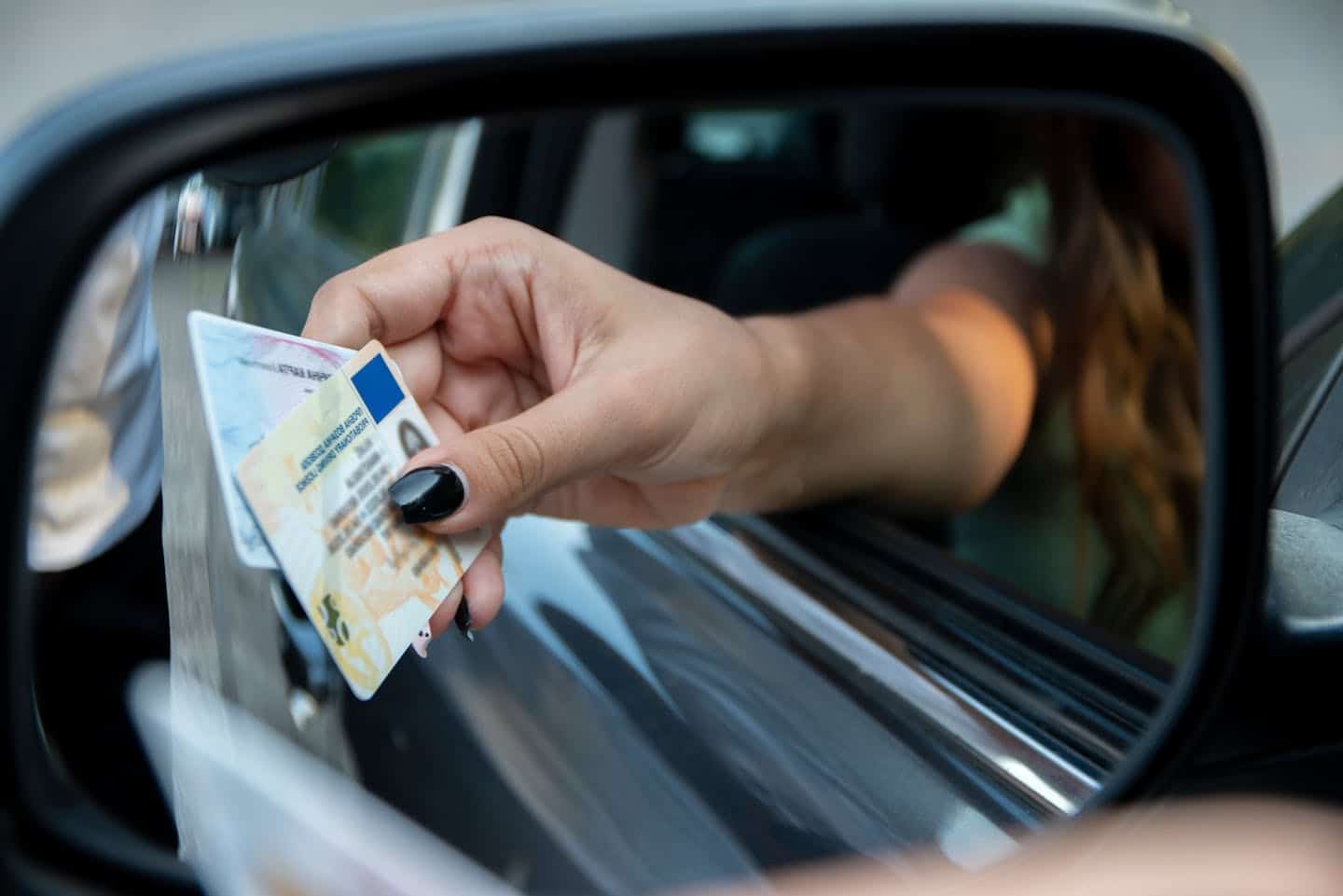 A person’s hand extends an ID card through a car's side mirror, suggesting a checkpoint or identification process, framed by the vehicle's window and exterior.