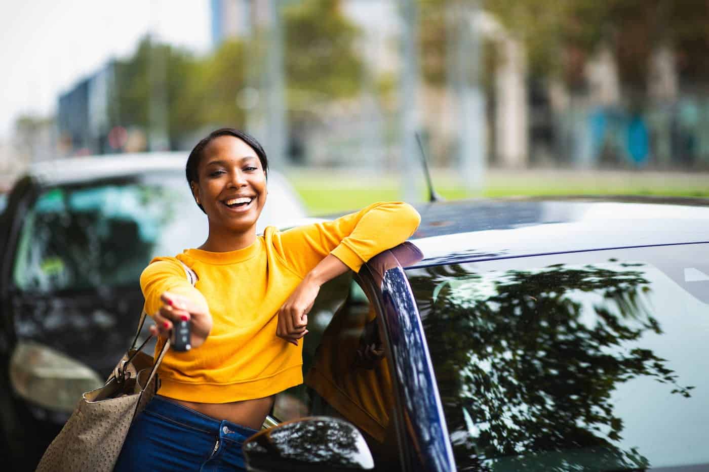 A smiling woman leans on a car, holding car keys, conveying a sense of pride or new ownership, in an urban setting with trees and buildings in the background.
