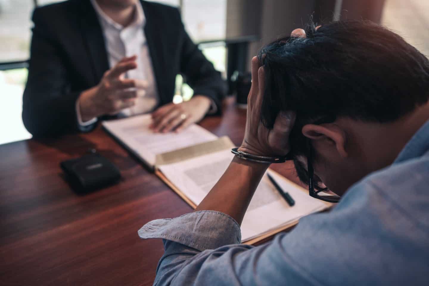 A person, head in hands, appears distressed during a meeting with another individual gesturing in explanation, set in an office environment with documents on the table.