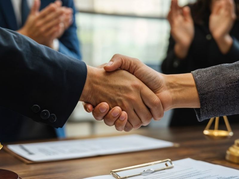 Two hands shake firmly, symbolizing agreement or partnership, over a wooden desk adorned with legal scales and documents, while others in the background applaud the gesture.