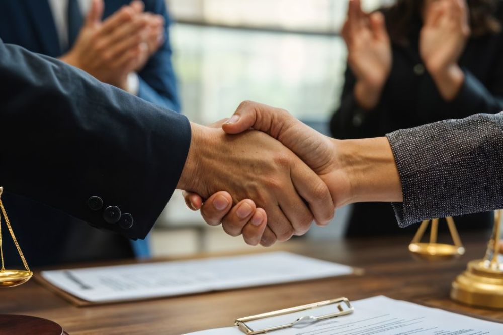 Two hands shake firmly, symbolizing agreement or partnership, over a wooden desk adorned with legal scales and documents, while others in the background applaud the gesture.