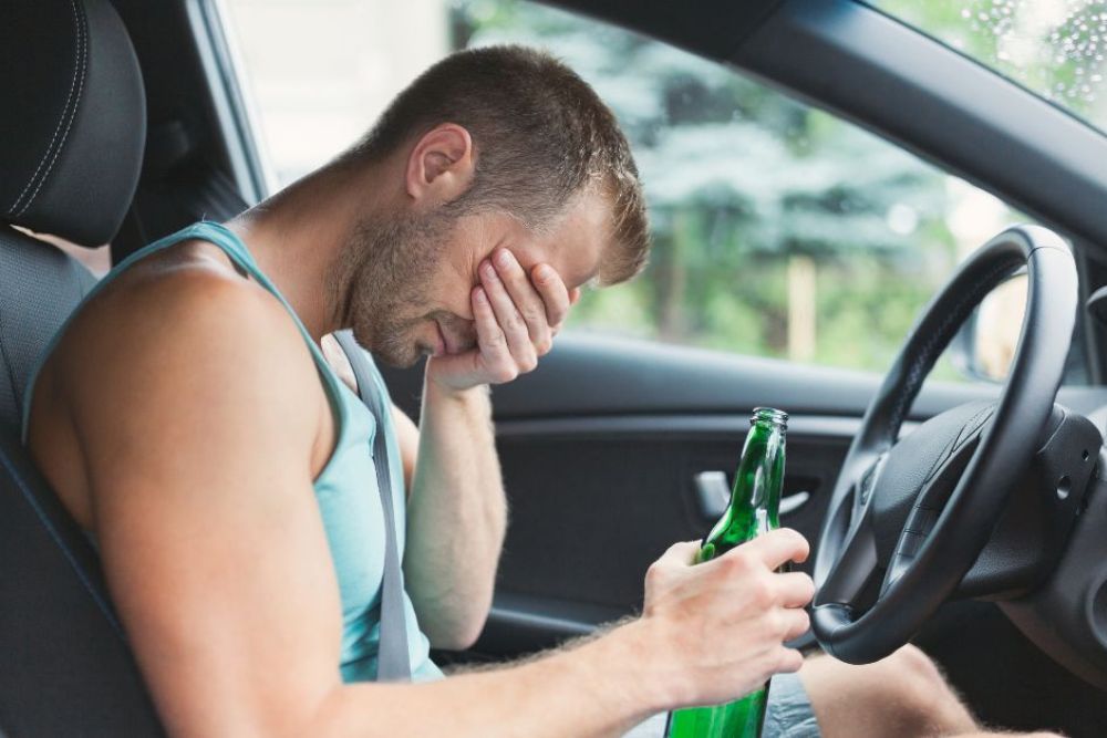 A man sits in a car, holding a green bottle and covering his face with one hand, showing signs of distress. The environment appears rainy with blurred greenery outside.