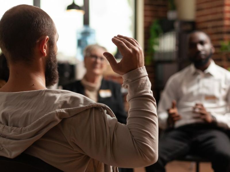 A man with short hair and a beard raises his hand while seated in a group discussion. Others listen attentively in a warmly lit, brick-walled room.