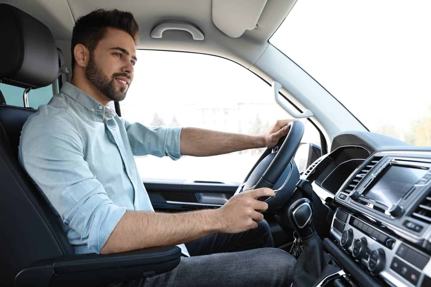 A man with a beard, driving a car, hands on the steering wheel, appears focused on the road ahead, inside a vehicle with a prominent dashboard.