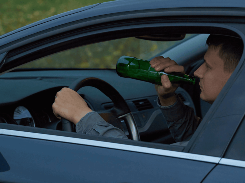 Driver drinks from a green bottle while steering a car, visible through the open window. The car interior is dark, and the outside shows green foliage.