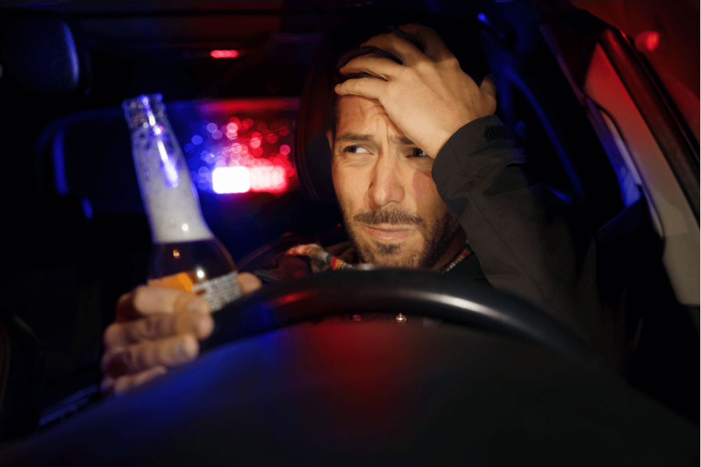 man holding a beer behind the wheel of car with police lights behind him