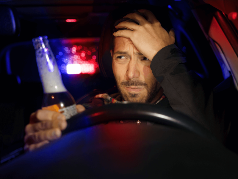 man holding a beer behind the wheel of car with police lights behind him