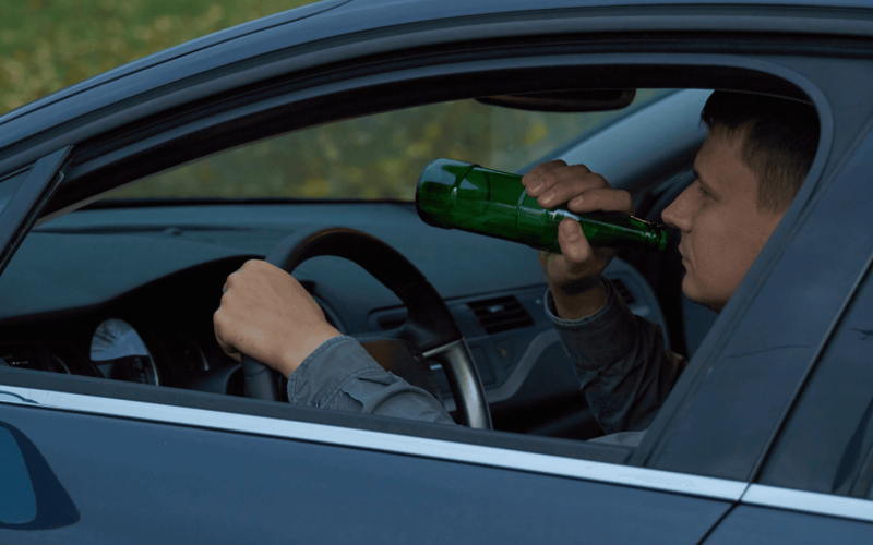 Driver drinks from a green bottle while steering a car, visible through the open window. The car interior is dark, and the outside shows green foliage.