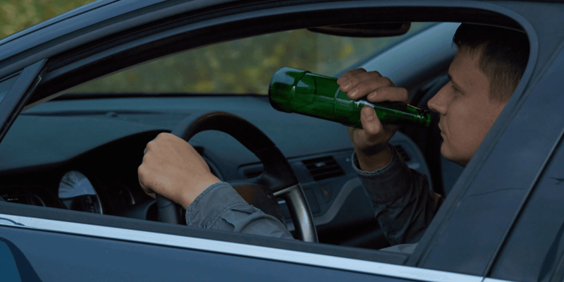 Driver drinks from a green bottle while steering a car, visible through the open window. The car interior is dark, and the outside shows green foliage.