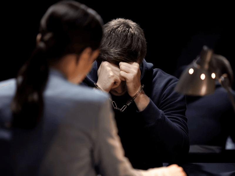 A person is sitting with their head in their hands, visibly stressed. Another blurred individual is in the foreground, at a desk under a lamp, indicating a competitive or examination setting.