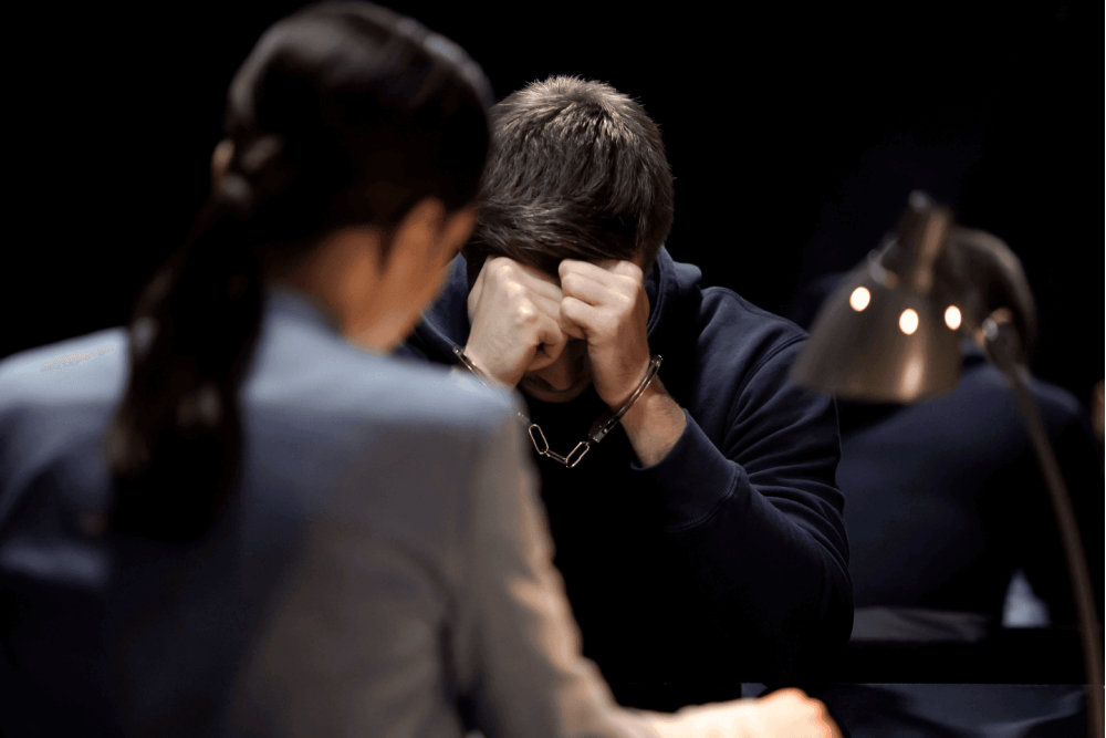 A person is sitting with their head in their hands, visibly stressed. Another blurred individual is in the foreground, at a desk under a lamp, indicating a competitive or examination setting.