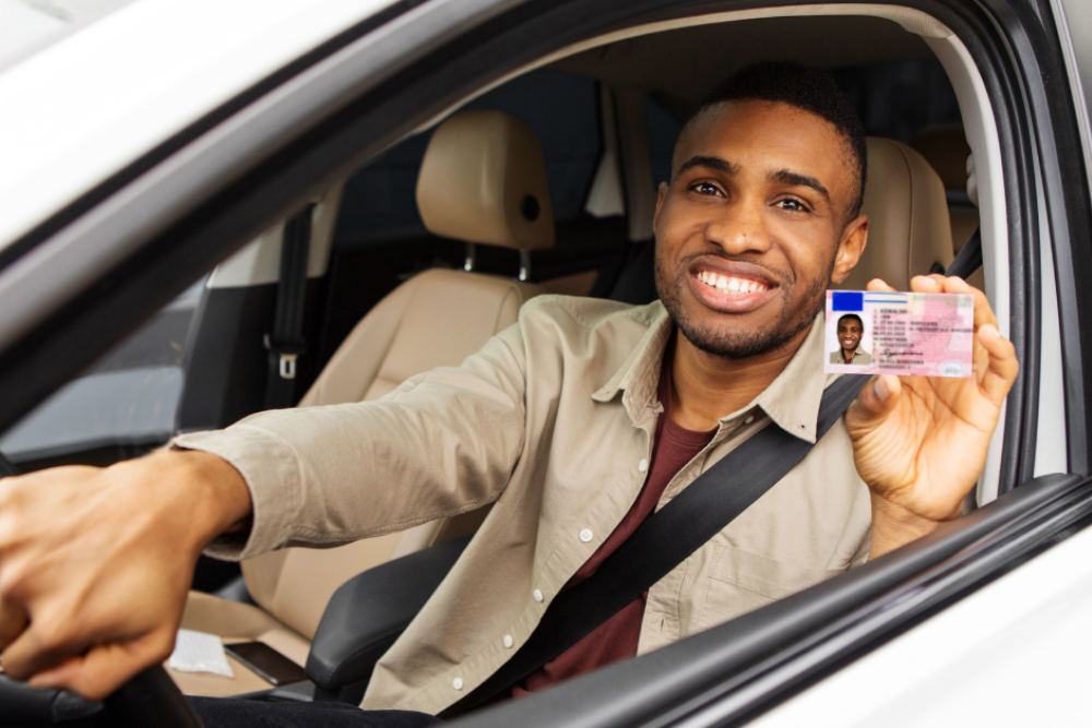 A young man holding a driver's license smiles while sitting in the driver's seat of a car. The interior features beige seats, and a steering wheel is visible in his hand.